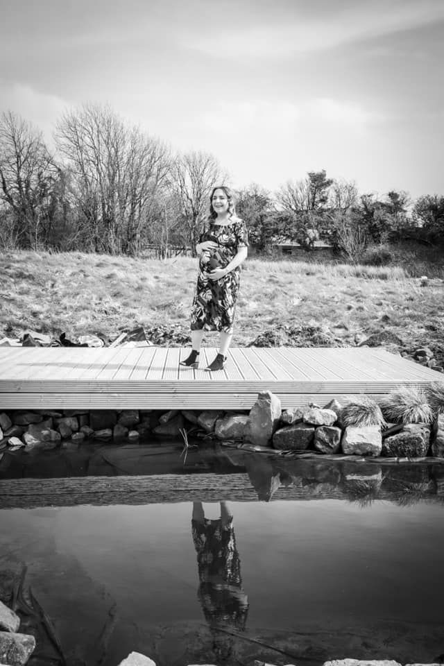 Black and white photo of a person standing on a wooden dock with their reflection in still water below.