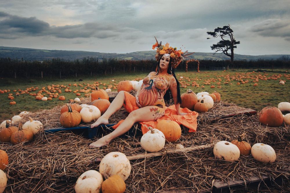 Creative autumn maternity photo with pumpkins and floral antler crown in field at sunset.