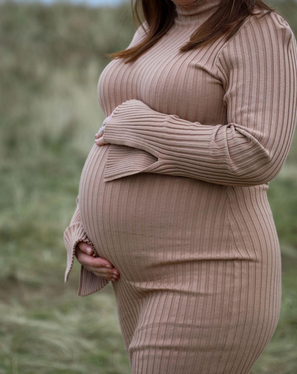 Close up of a pregnant belly in a beige ribbed sweater dress against a natural outdoor background.