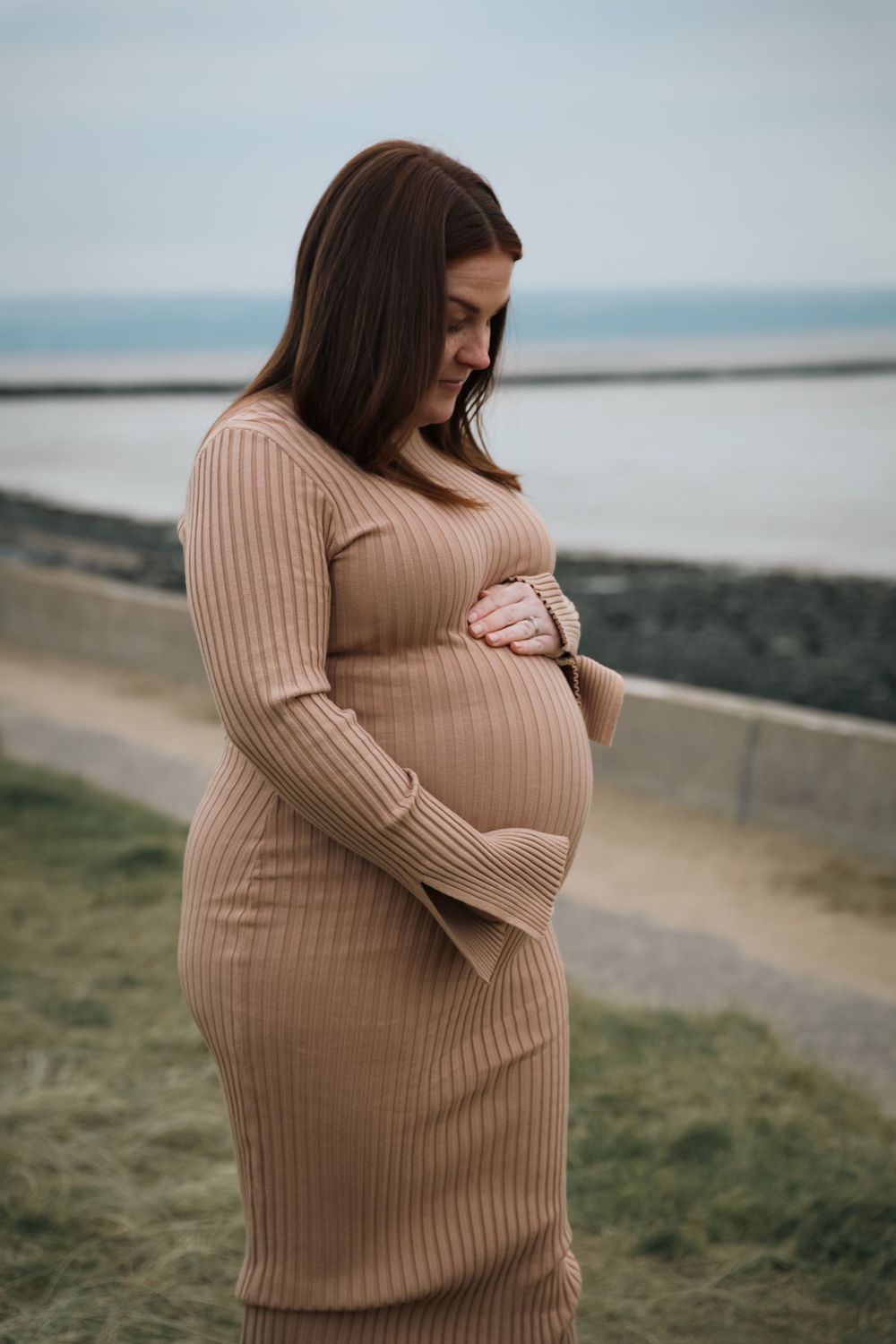 Expecting mother in beige ribbed dress gazes down at her baby bump near coastal setting.