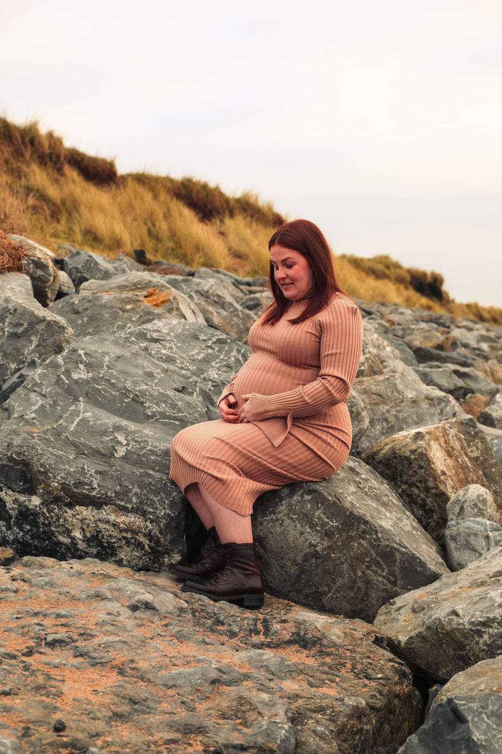 Pregnant woman in beige sweater dress sits on coastal rocks during sunset maternity shoot.