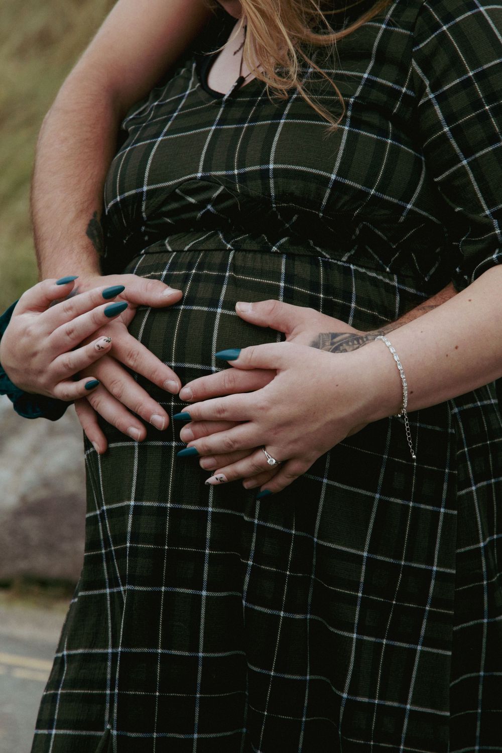 Close-up of hands gently resting on pregnant belly wearing plaid dress with blue nail polish.