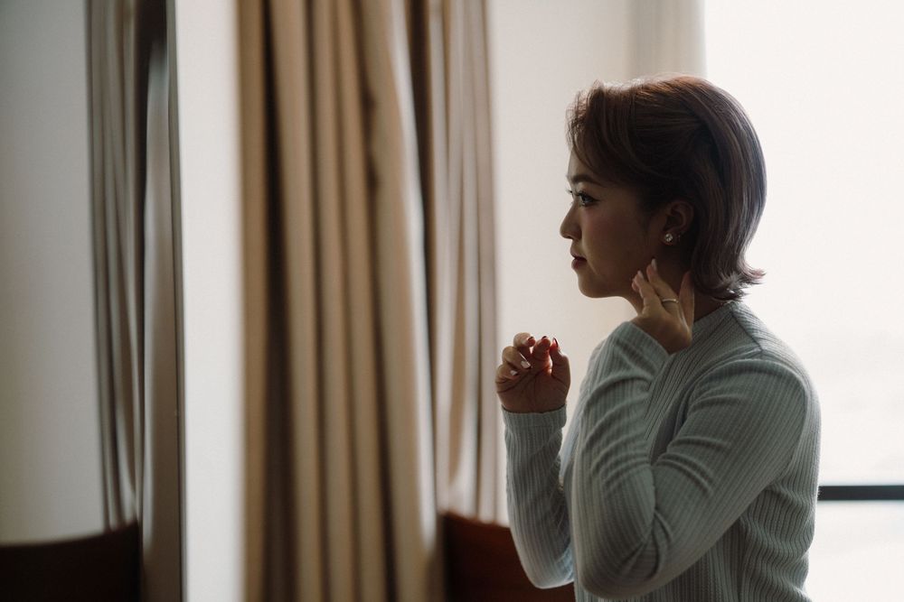 A person sits alone near a window with hands clasped, looking contemplative in a dimly lit room.