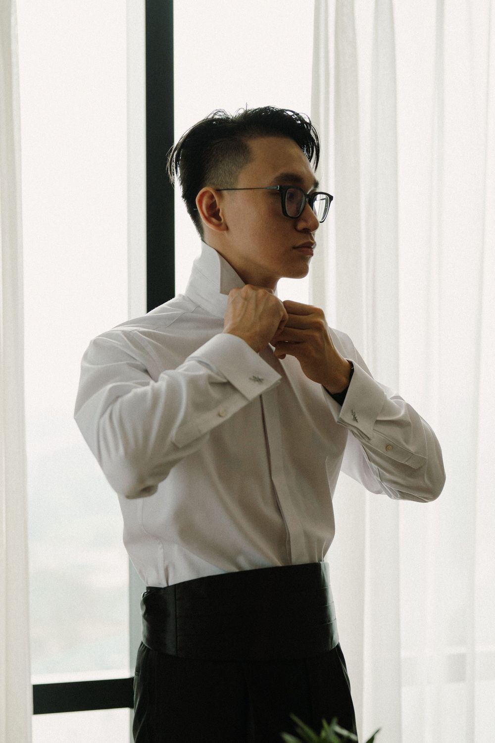 A person in a white shirt adjusts their glasses while standing by window curtains in contemplative pose.