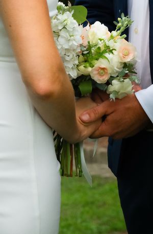 A couple holds hands during a wedding ceremony with a beautiful bouquet of white hydrangeas and peach roses.