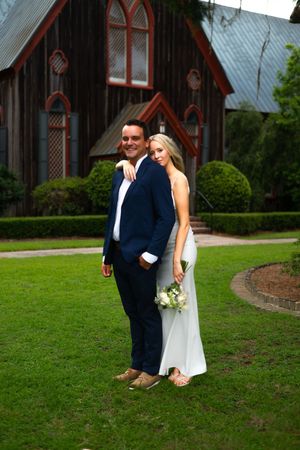 A wedding couple poses on a lawn in front of a charming gothic-style church with arched windows.