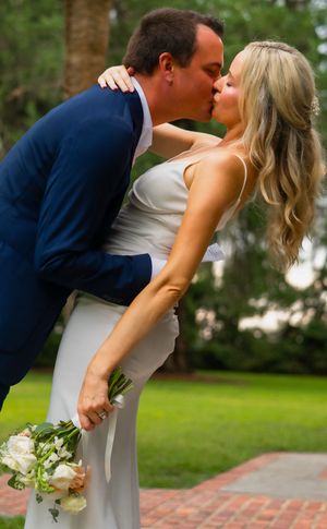 A romantic wedding kiss between newlyweds in formal attire, with the bride holding a white bouquet in an outdoor garden setting.