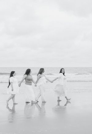 A group in white outfits walks along a misty beach holding hands in a joyful line formation.
