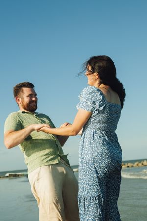 A couple in casual attire dances and laughs together on a sunny beach with blue ocean waters in the background.