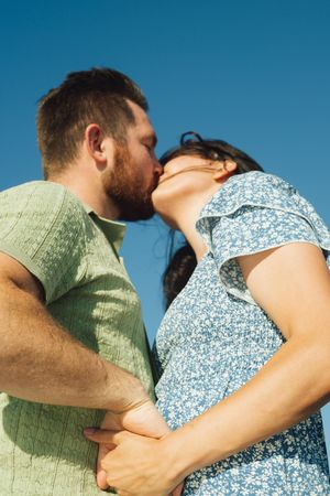A romantic couple in a blue and green outfits embrace and kiss against a bright blue sky background.