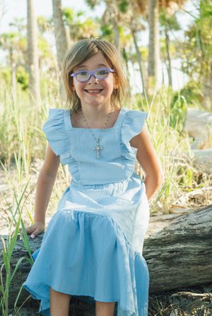 Young child wearing glasses and a light blue ruffled dress poses outdoors among palm trees and natural scenery.