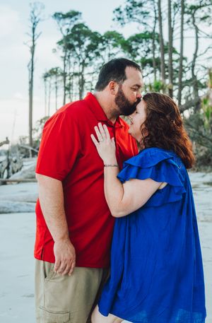 A couple in red and blue outfits share an intimate moment on a beach with tall pine trees in the background.