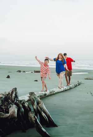 Three people balance and play on a large driftwood log at the beach with waves and rocks in the background.