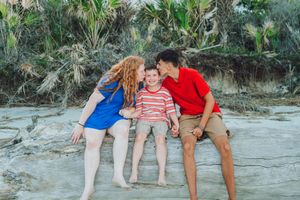 Three people sit together and smile while relaxing on a sandy beach near tropical foliage.