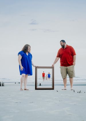 Creative beach photo composition with couple holding picture frame showing smaller figures walking on sandy shore.