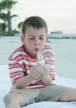 Young child in red and white striped shirt sitting on beach blowing a dandelion flower on sunny day.