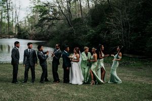 Wedding party in sage green dresses and dark suits pose playfully by a lake in a wooded setting.