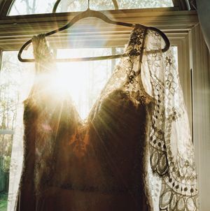 Vintage lace wedding dress hanging in sunlit window with delicate details and ethereal morning glow.