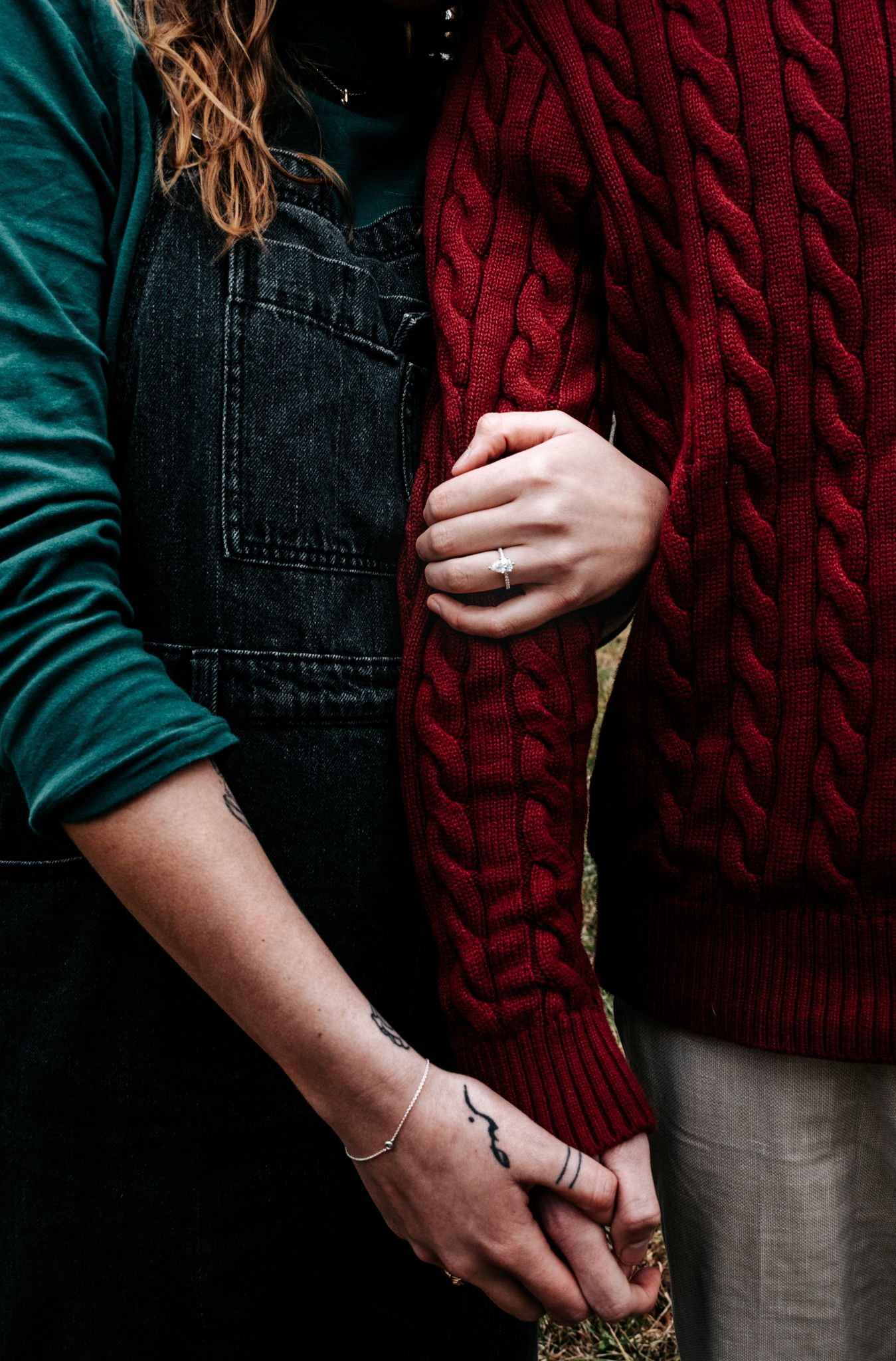 Close-up of two people holding hands, wearing denim overalls, green sleeve and red cable knit sweater.