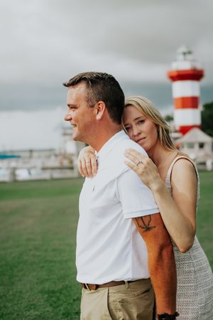 A romantic couple poses in front of a red and white lighthouse tower on a cloudy day by the coast.