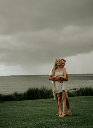 A couple embraces on a grassy hilltop overlooking a moody ocean view under stormy gray skies.
