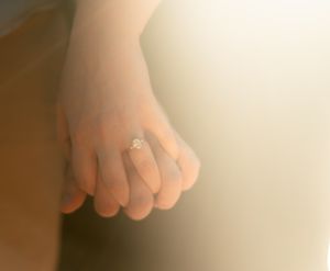 A close-up view of an engagement ring on a hand against a soft, blurred background.