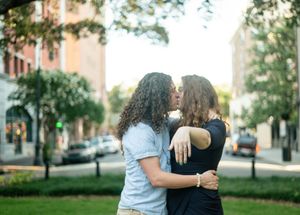 Two people in casual attire share a close moment together on a city street with buildings and trees in the background.