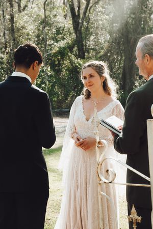 A romantic outdoor wedding ceremony takes place in a sunlit forest setting with rays of light streaming through the trees.