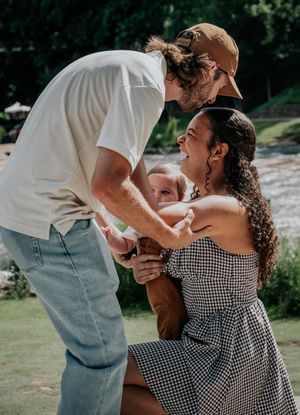 Two people share a tender moment while holding a baby outdoors on a sunny day.
