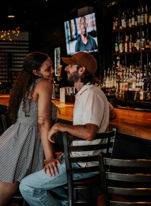 A couple shares an intimate moment at a dimly lit bar with a gingham dress and baseball cap.