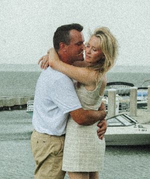 A romantic couple embraces while standing near a coastal marina on an overcast day.