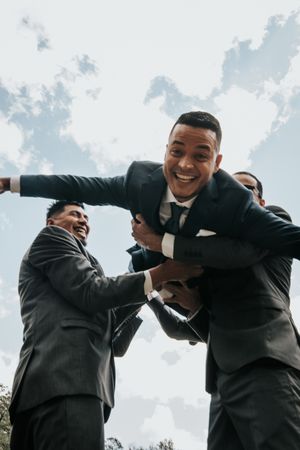 Groomsmen in suits lift and carry someone playfully during outdoor wedding celebrations against cloudy sky.