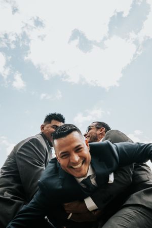 A group of people in dark suits share a lighthearted moment under a bright cloudy sky.