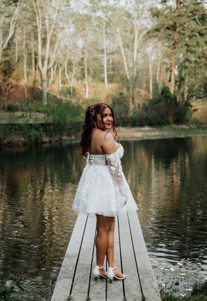 A woman in a short white lace wedding dress poses on a wooden dock by a serene lake surrounded by trees.
