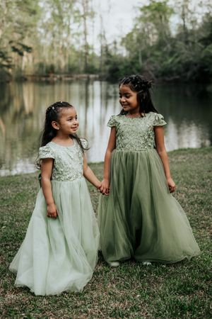 Two young girls in matching sage green lace and tulle gowns hold hands by a tranquil lake in a wooded setting.