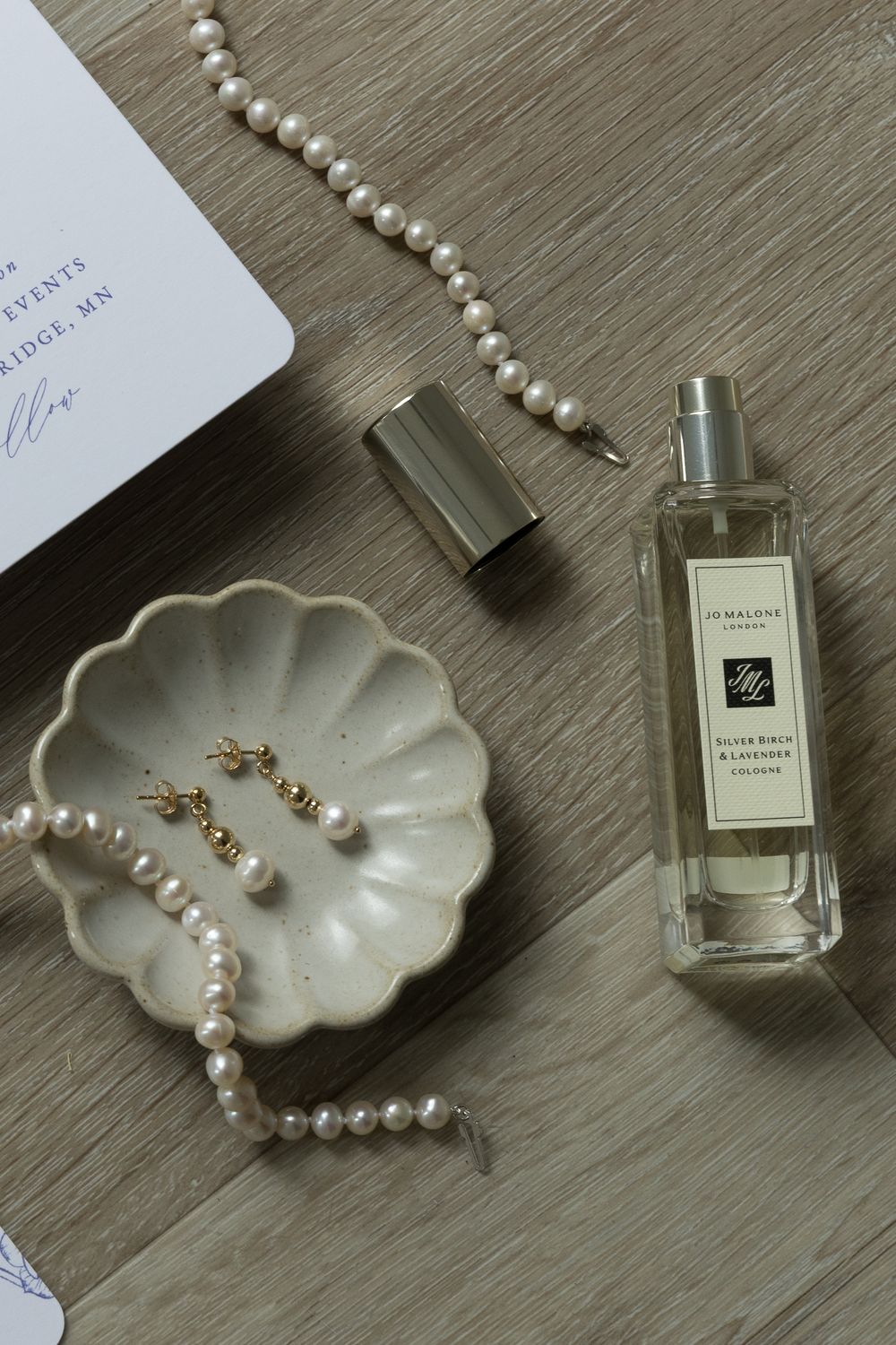Wedding accessories including pearl jewelry, perfume bottle and shell dish arranged on wooden surface.