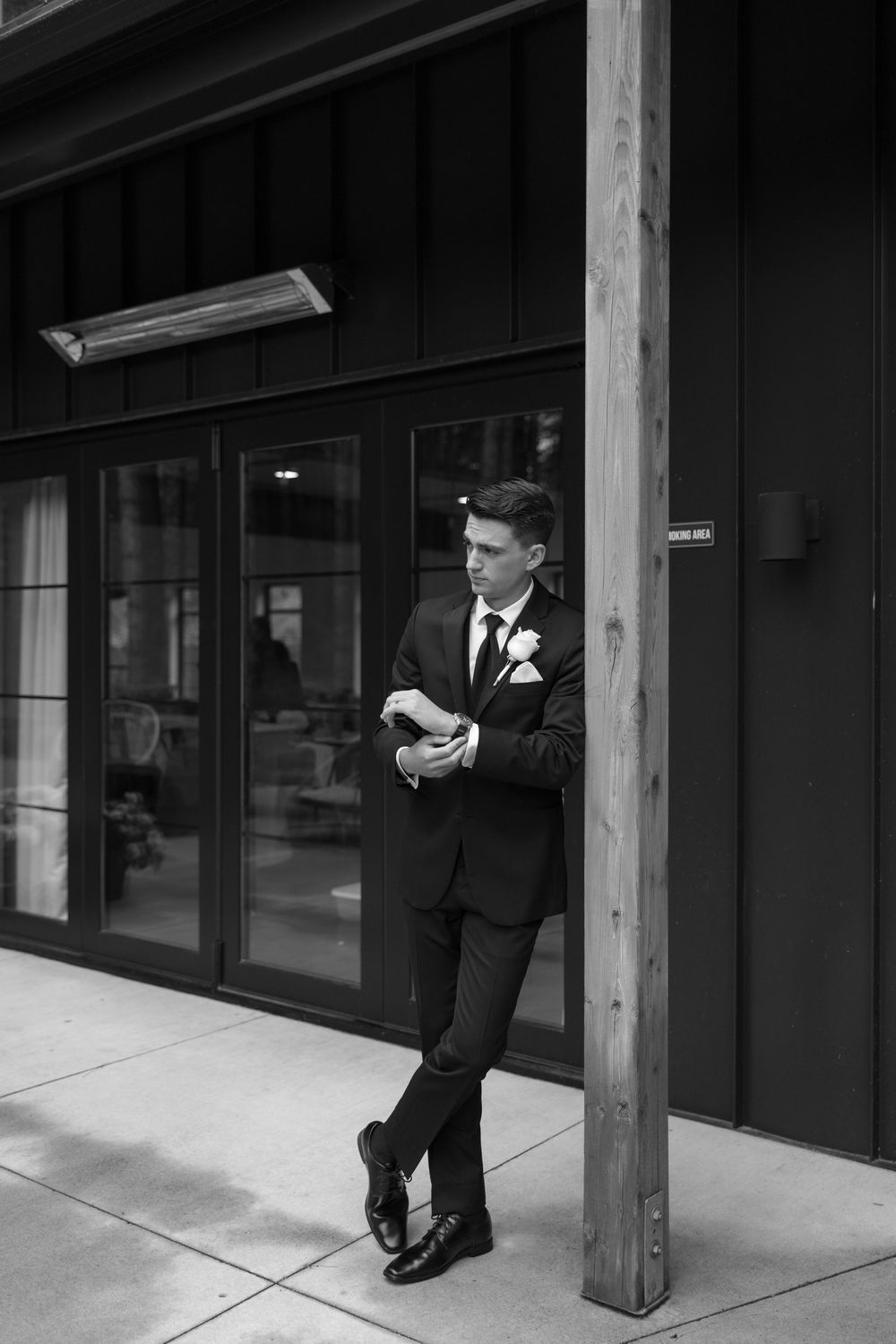 Groom in formal black suit waiting outside modern building in black and white photograph.