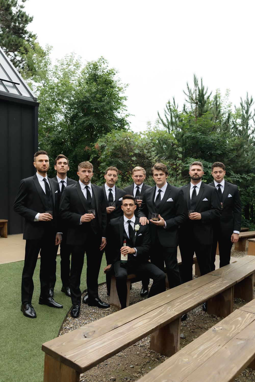 Groomsmen in black suits pose casually on wooden benches outdoors with drinks in hand.