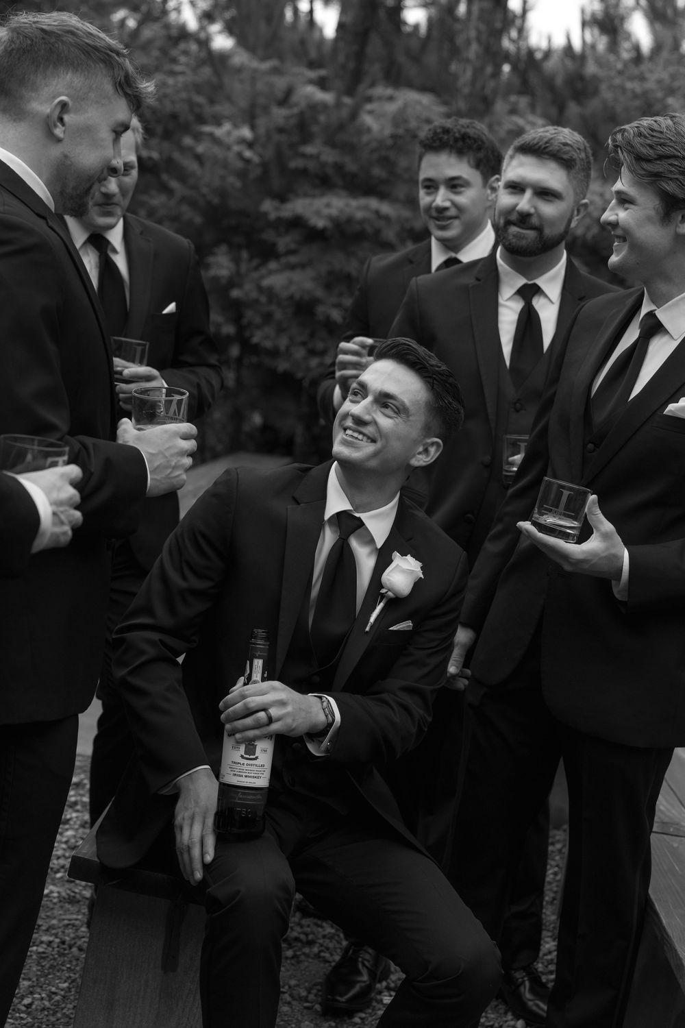 Groomsmen in dark suits sharing drinks and laughs at outdoor wedding celebration.