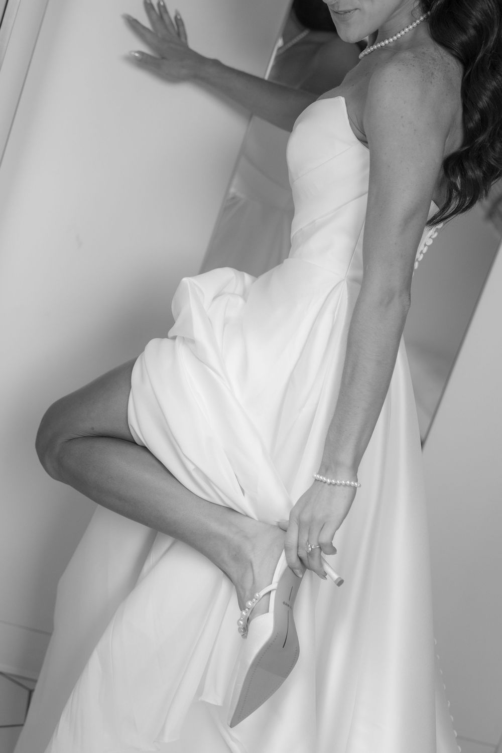 Black and white photo of bride lifting wedding dress to adjust shoe strap.