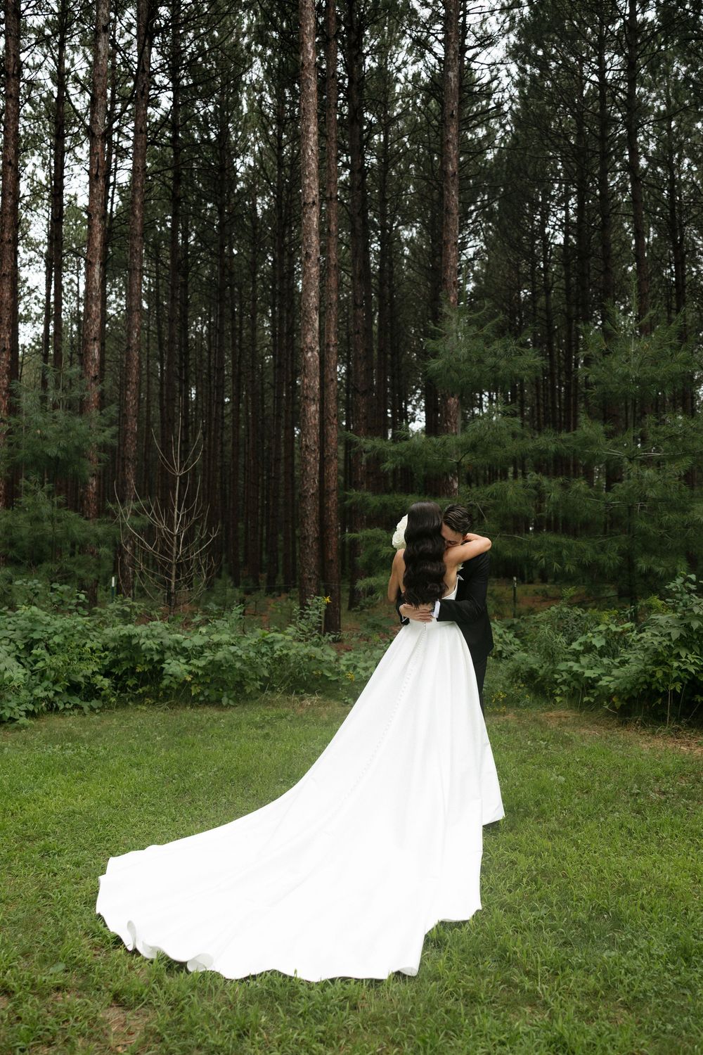A bride in a flowing white gown with a dramatic train stands in a serene pine forest setting.