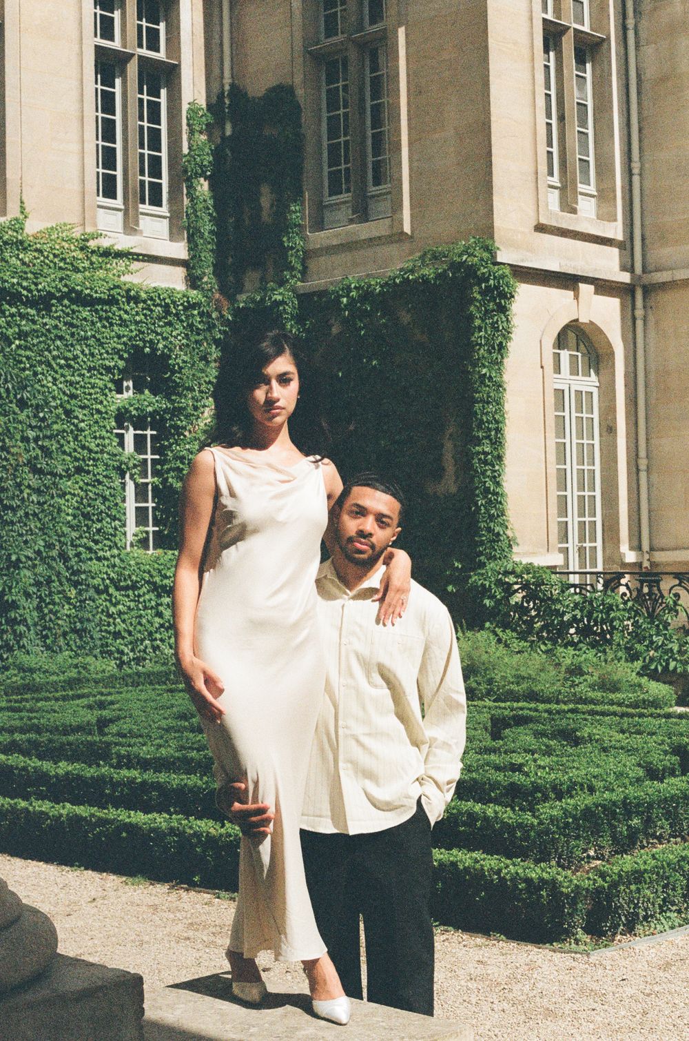 A couple poses in front of an ivy-covered historic building with manicured hedges in four candid shots.
