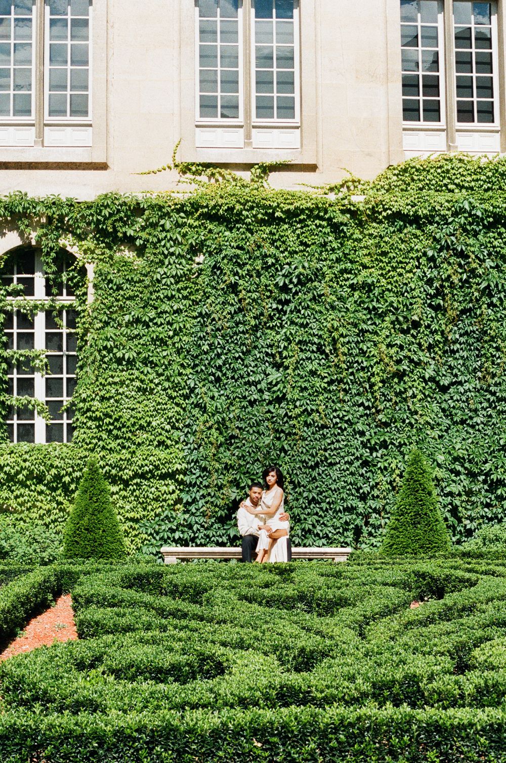 A pair sit together on a stone bench surrounded by perfectly manicured green hedges and ivy-covered walls.