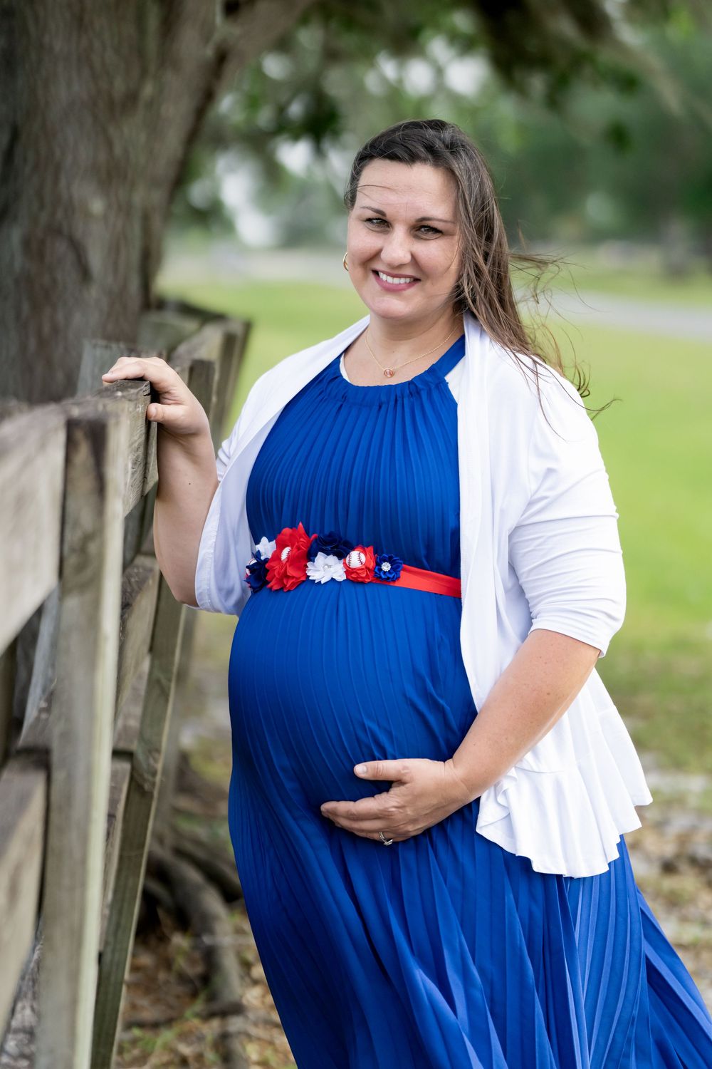 Outdoor maternity photos by a wooden fence featuring a blue dress with red and white floral accents.