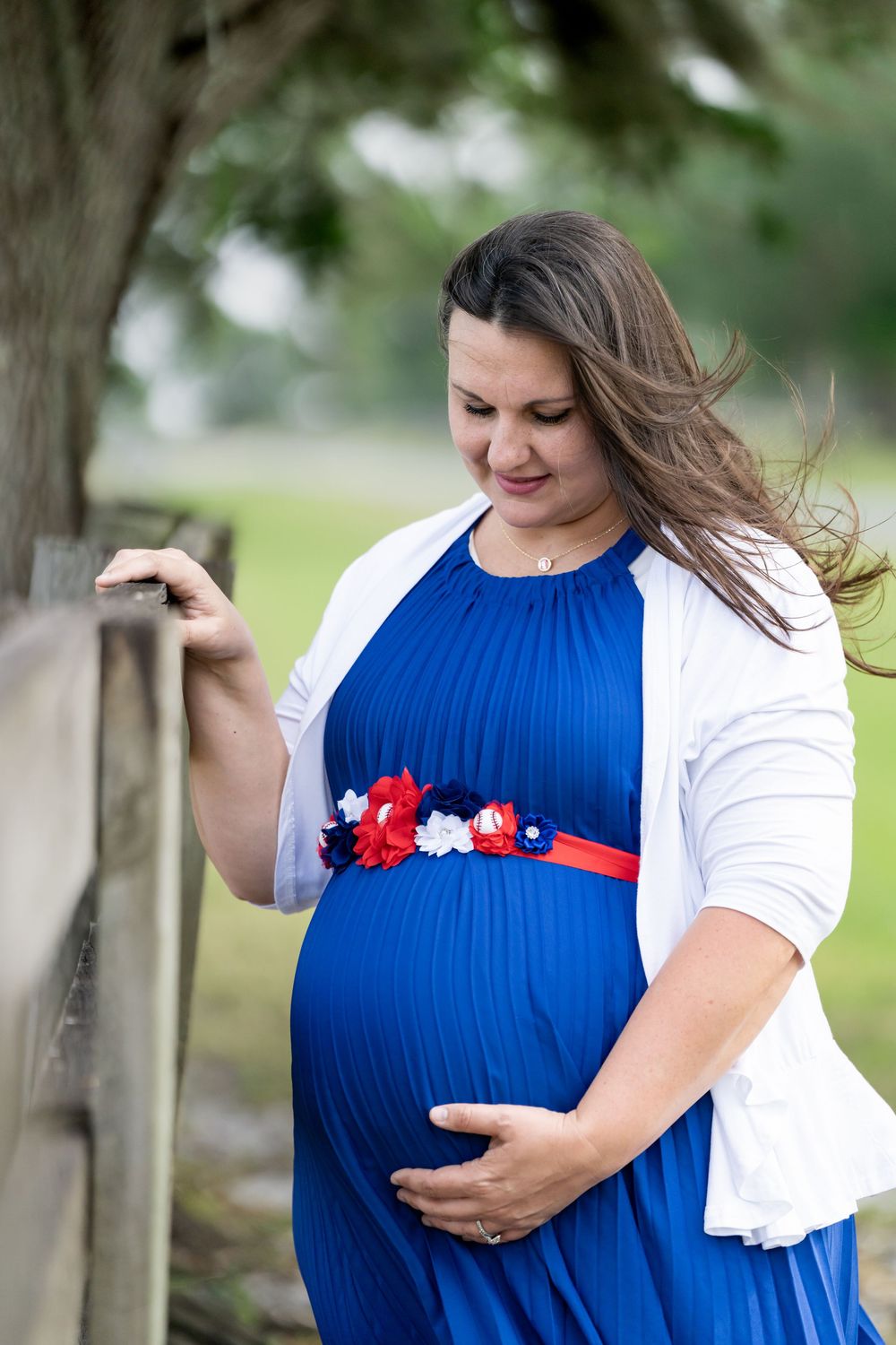 Maternity portraits in a blue dress against a natural outdoor backdrop with wooden fencing.
