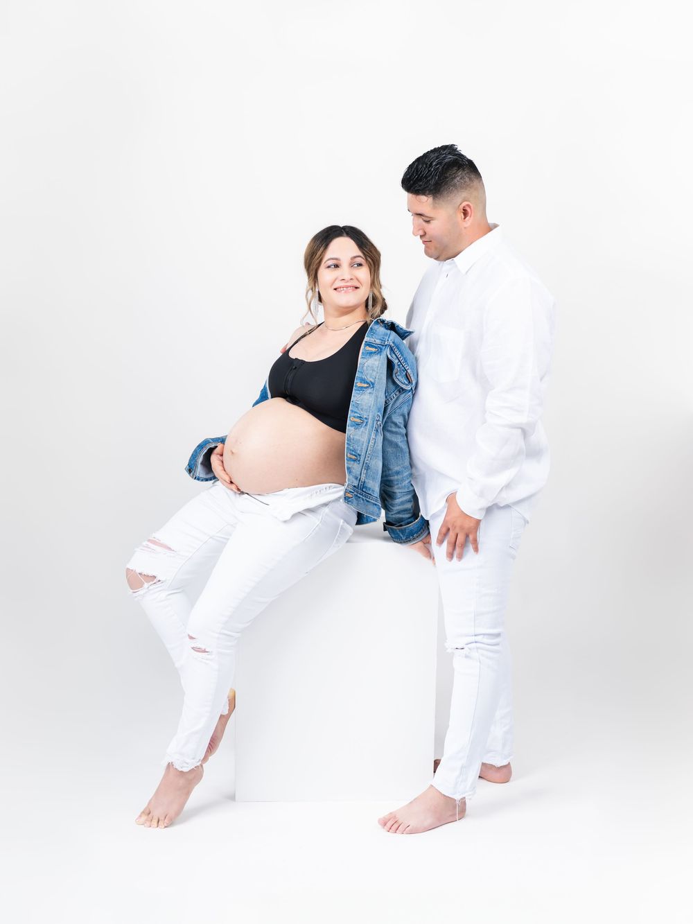 A maternity photoshoot featuring a couple in white and denim clothing against a white background.