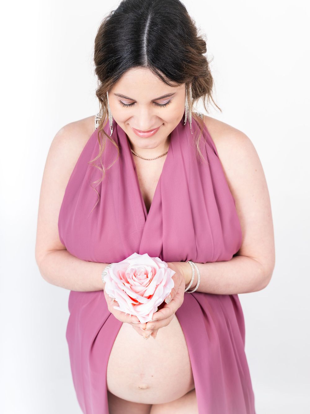 A pregnant woman in a pink dress holds a delicate rose while looking down at her belly.