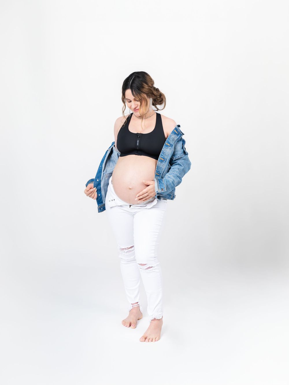 Casual maternity photos with denim jacket and white pants in bright studio setting.
