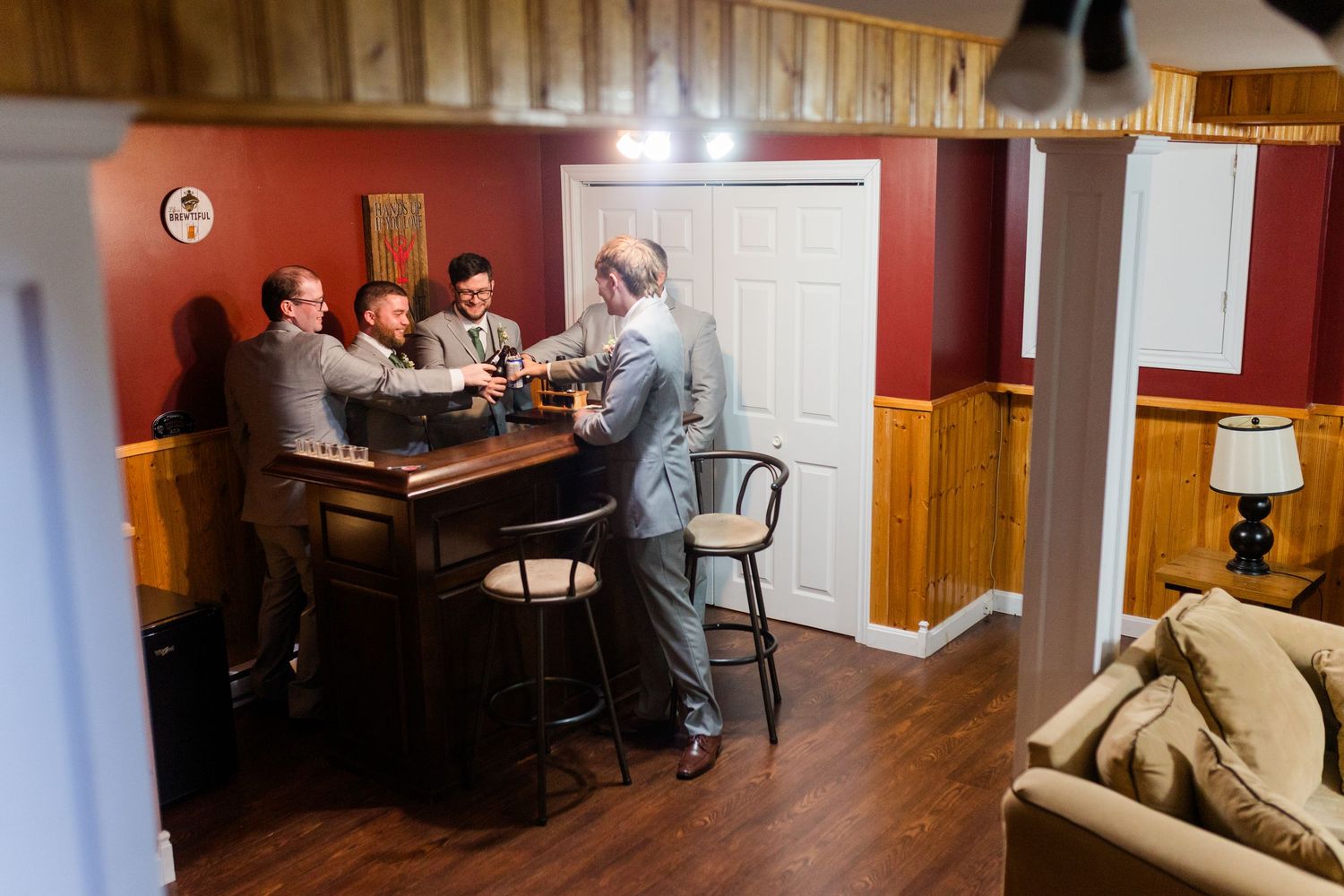 Wedding party gathers around a wooden bar in rustic basement with red walls and wood wainscoting.