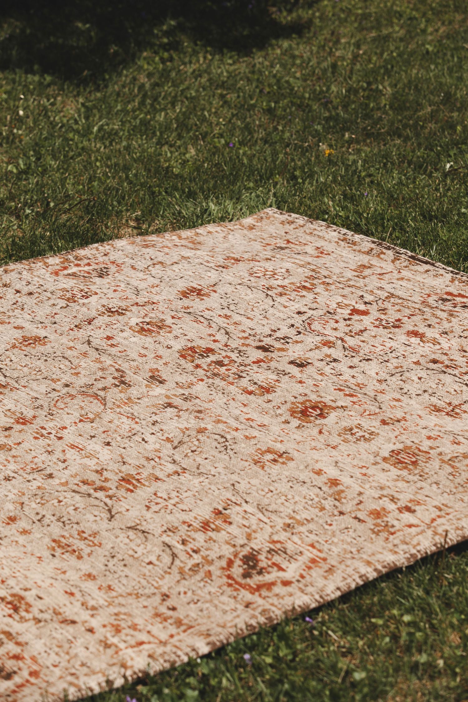 A vintage distressed area rug with rust-colored patterns lies on green grass in an outdoor setting.